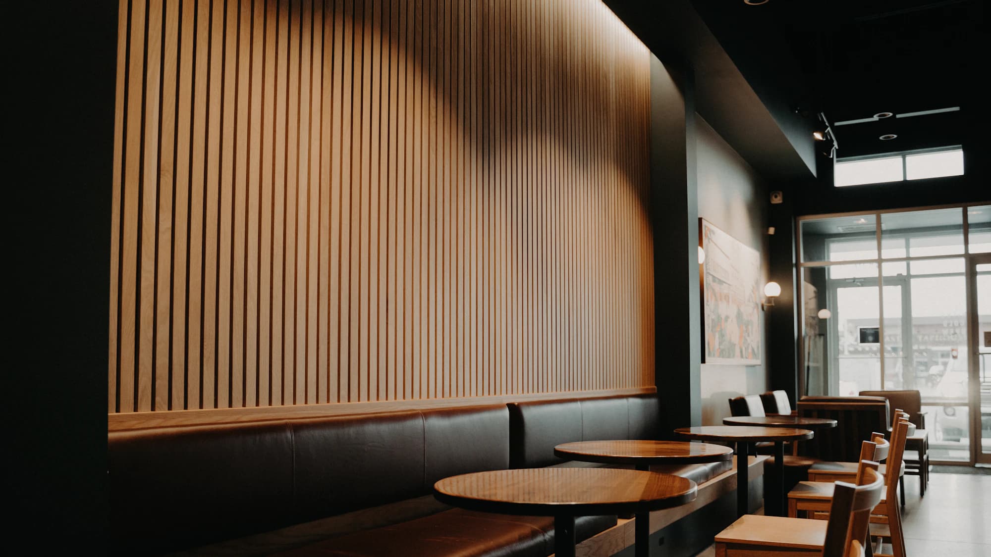 Round wooden tables along a slatted timber wall, with daylight coming in from windows at the far end.