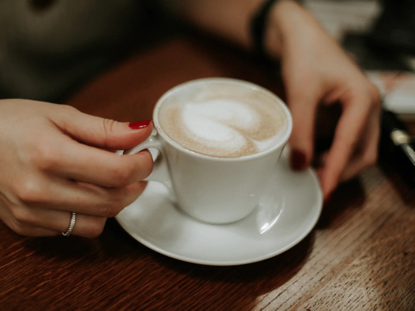 A pair of hands resting around a white cup of coffee on its saucer, on a dark wooden counter.
